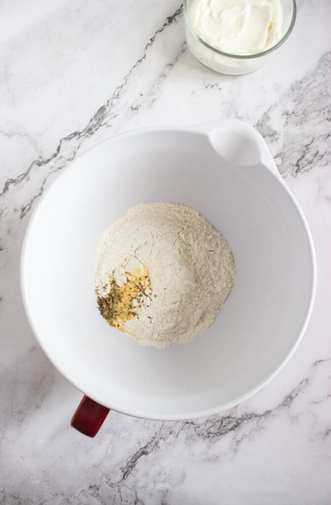 A white mixing bowl with flour and spices sits on a marble countertop; a small glass bowl of sour cream is nearby.
