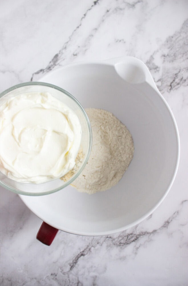 A glass bowl of yogurt being poured into a white mixing bowl containing flour, set on a marble countertop.