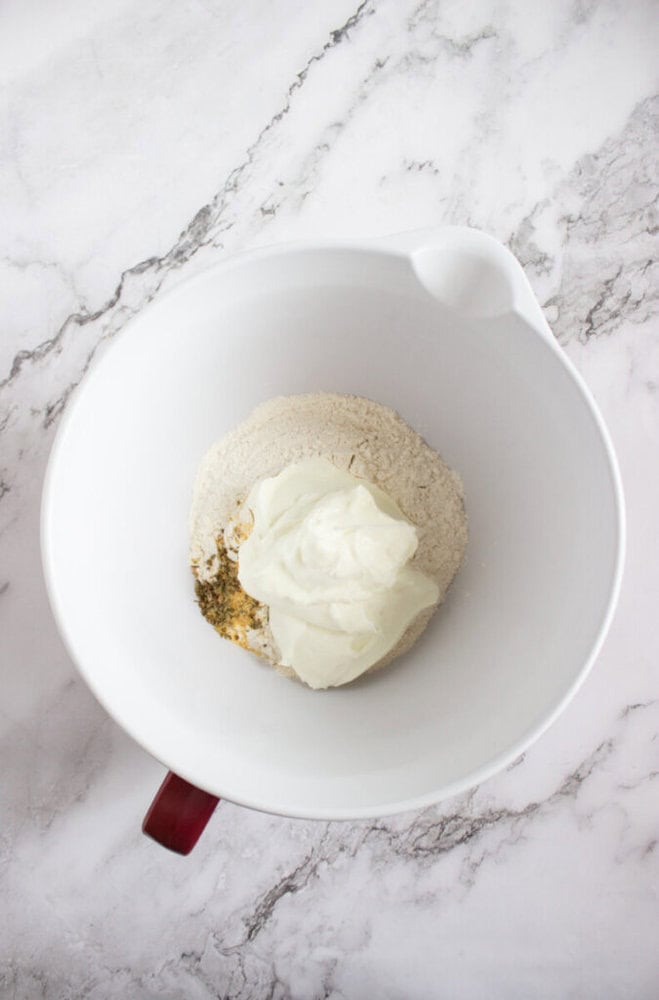 A white mixing bowl on a marble surface contains flour, a dollop of sour cream, and various spices.