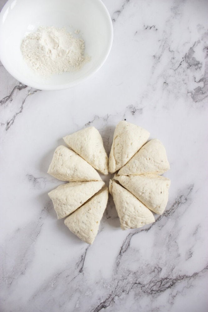 Eight pieces of unbaked biscuit dough arranged in a circle on a marble surface, with a white bowl containing flour in the upper left corner.