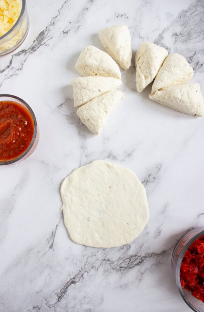 Raw pizza dough divided into six pieces, one piece rolled flat, with bowls of tomato sauce and shredded cheese on a marble surface.