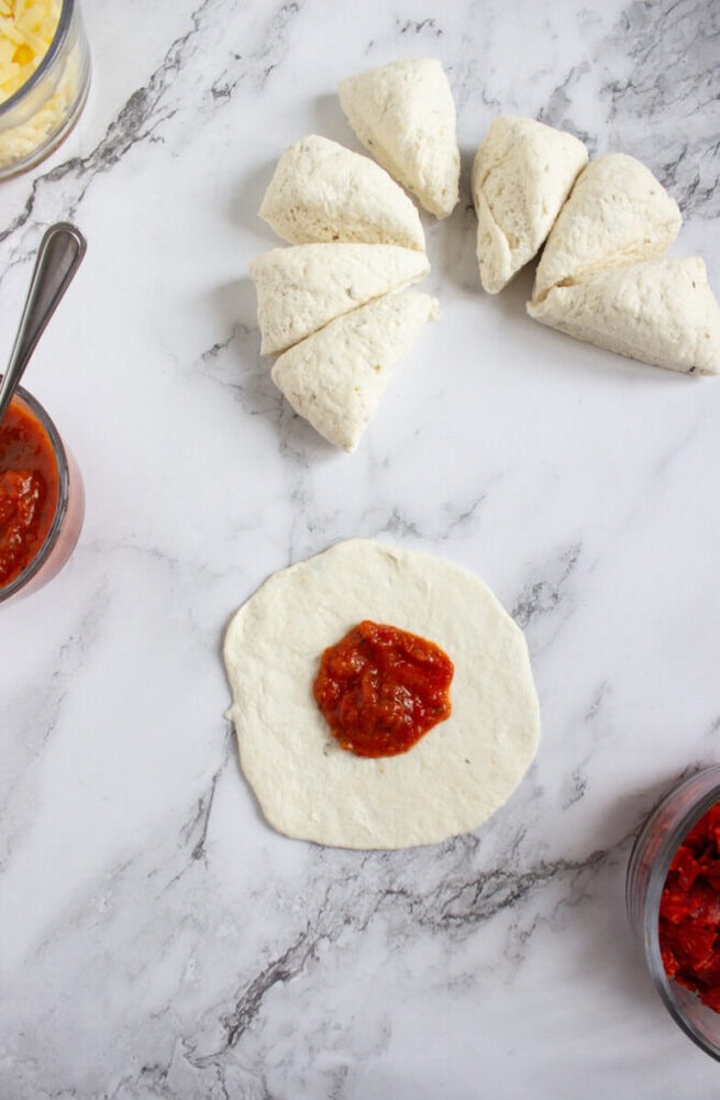 Pizza dough divided into pieces, one piece rolled flat with tomato sauce in the center, surrounded by bowls of sauce on a marble surface.