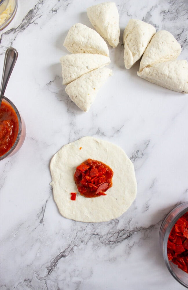 Pizza dough divided into pieces on a marble surface, with one piece rolled out and topped with tomato sauce. Bowls of pizza ingredients are nearby.