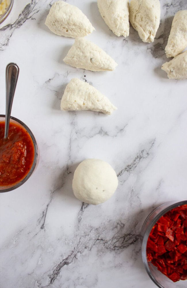 Dough pieces and a dough ball on a marble surface, with bowls of tomato sauce and sliced pepperoni nearby.