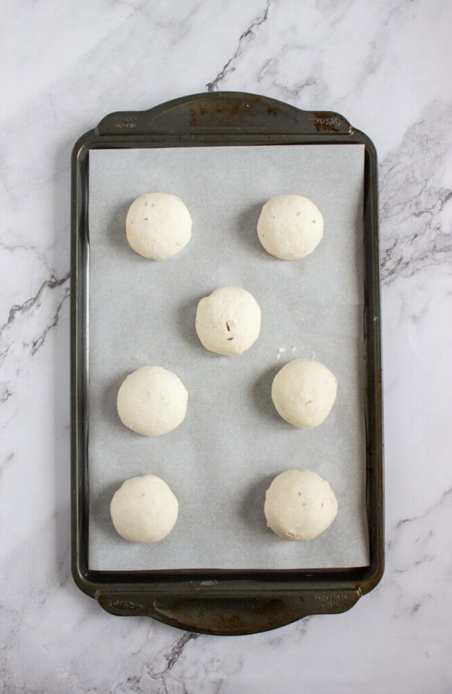 Six balls of dough are evenly spaced on a parchment-lined baking sheet, placed on a marble countertop.