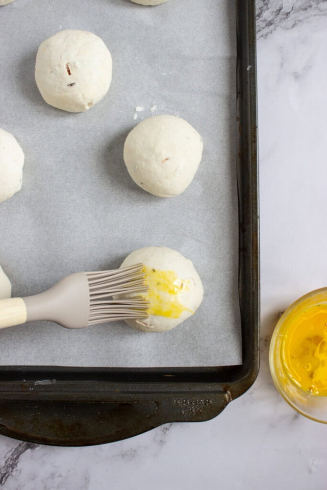 Unbaked dough balls on a parchment-lined baking sheet, with one being brushed with egg wash using a pastry brush; a bowl of beaten egg is nearby.