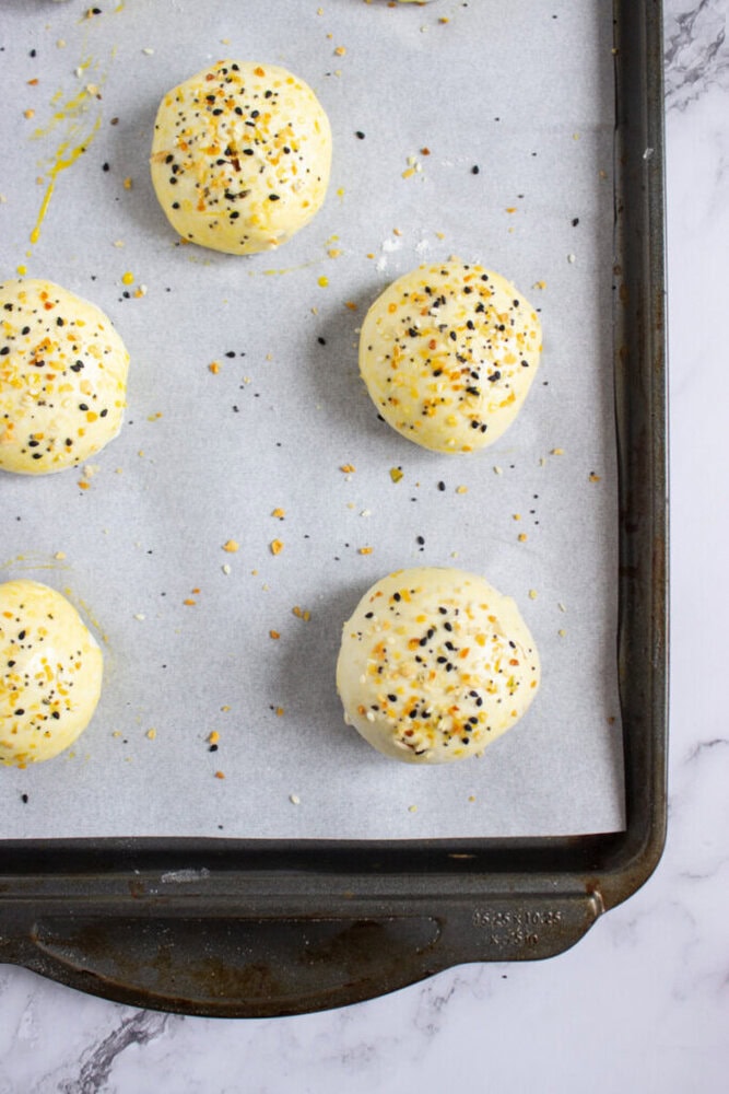 Six unbaked dough balls topped with seeds and seasoning are arranged on a parchment-lined baking sheet.