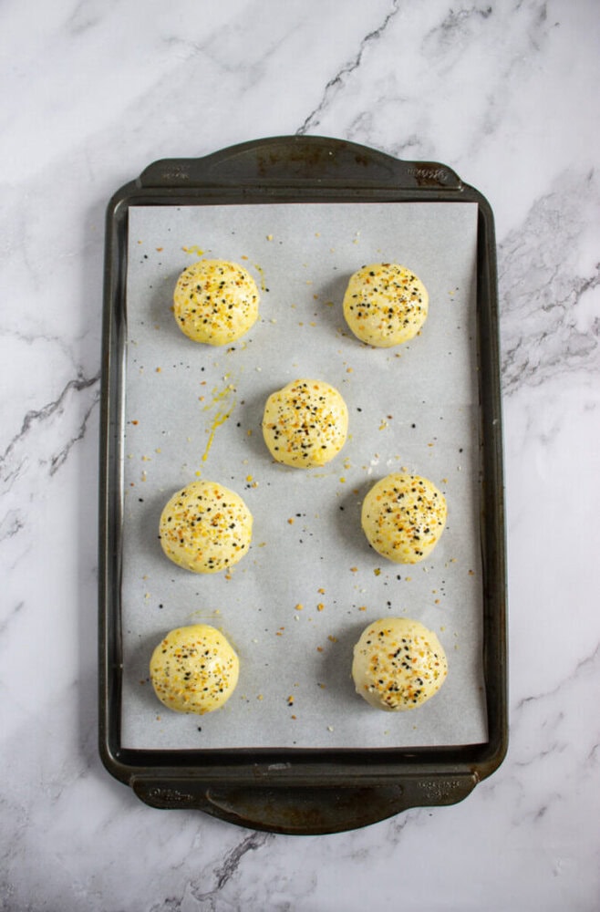 A baking tray with seven round, unbaked dough balls topped with seasoning, placed on parchment paper over a marble surface.