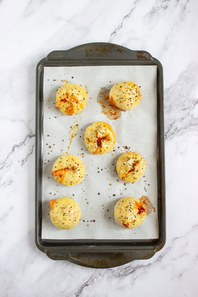 A baking tray with seven round, golden-brown baked rolls topped with seeds and cheese, placed on parchment paper over a marble surface.