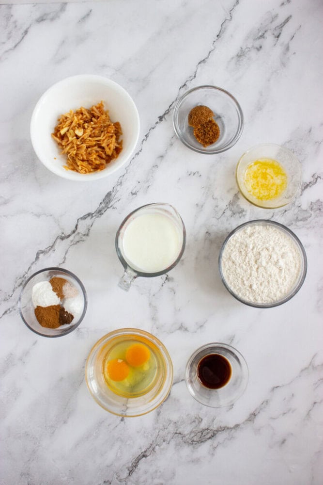 Various baking ingredients including flour, eggs, milk, vanilla extract, spices, melted butter, and shredded apples arranged in small bowls on a marble surface.