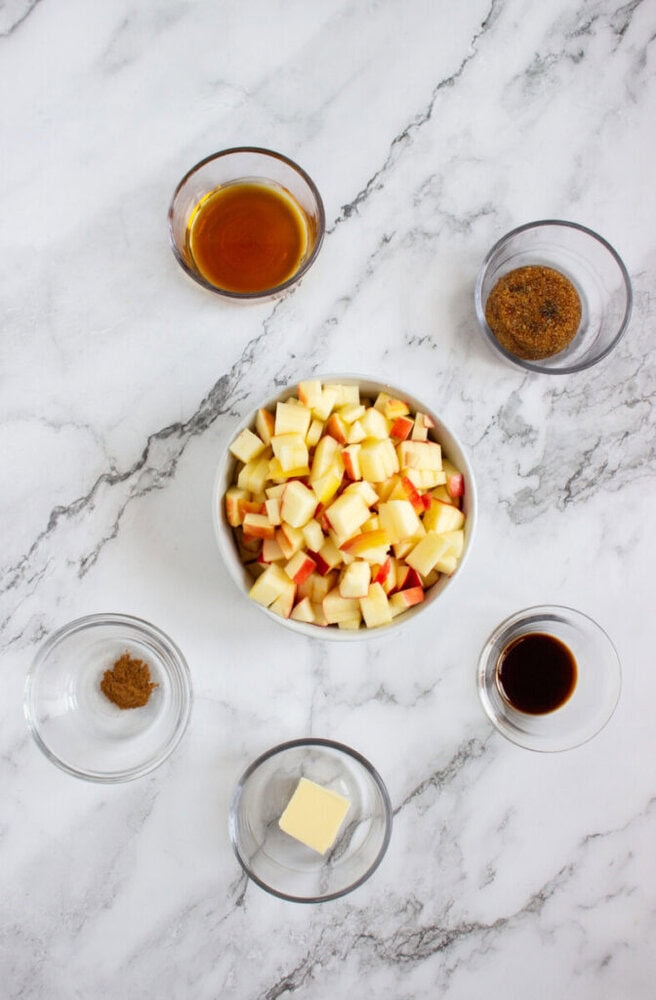 A bowl of chopped apples is surrounded by small glass bowls containing maple syrup, brown sugar, ground cinnamon, a pat of butter, and vanilla extract on a marble surface.