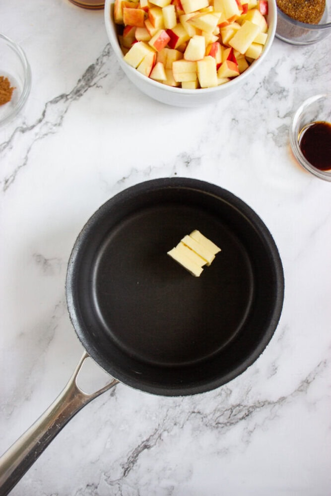 A small stack of butter slices in a black saucepan on a marble countertop, with bowls of chopped apples and other ingredients nearby.