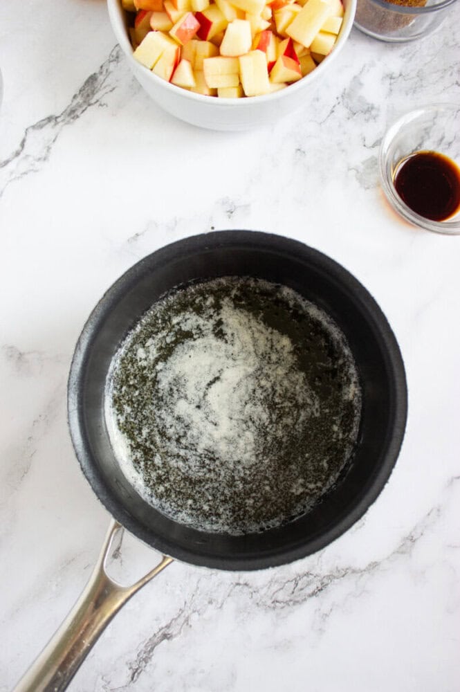 A saucepan with melted butter sits on a marble countertop, next to a bowl of diced apples and a small glass bowl of vanilla extract.