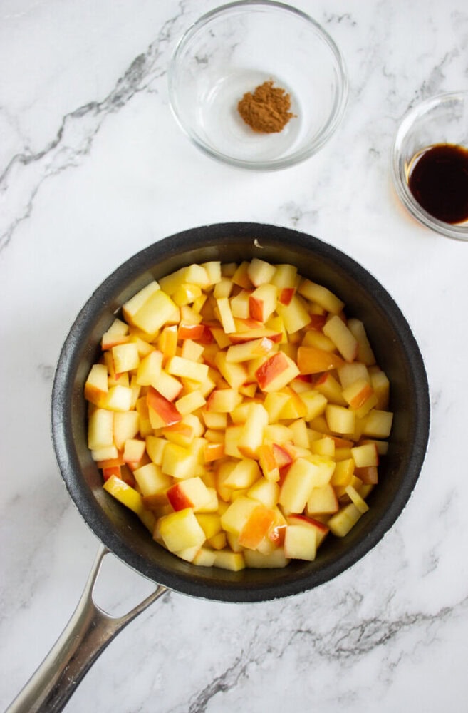 A saucepan filled with chopped apples sits on a marble surface, with small bowls of cinnamon and vanilla extract nearby.