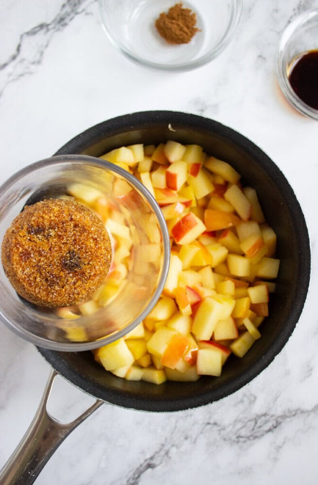 A saucepan filled with chopped apples, with a bowl of brown sugar being poured in; other ingredients in small bowls are nearby on a marble countertop.