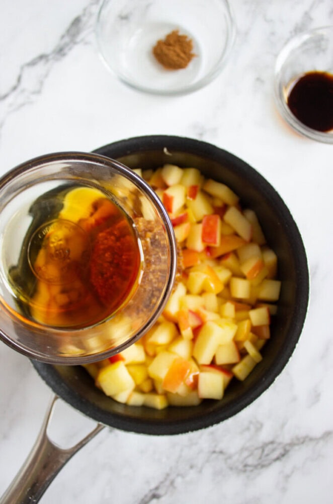 A saucepan with chopped apples, a bowl of liquid being poured in, and two small bowls with spices and vanilla extract on a marble countertop.