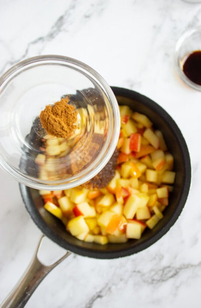 A glass bowl of ground cinnamon is being added to a saucepan with chopped apples on a marble countertop.