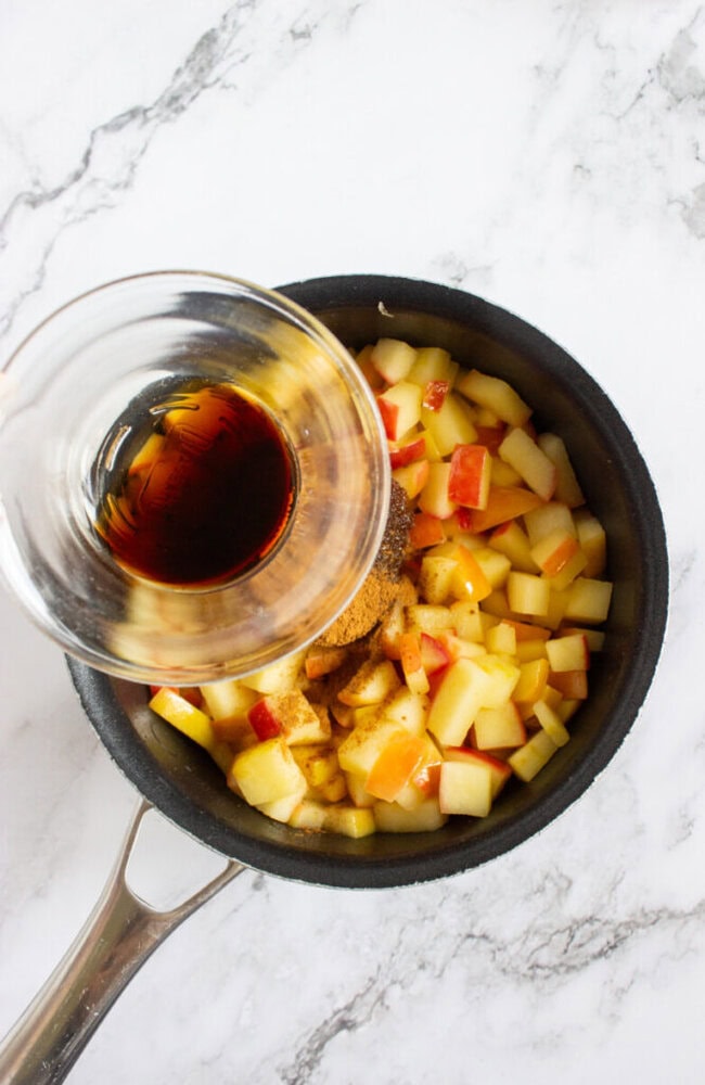 A saucepan with chopped apples and spices, with a glass bowl of liquid being poured in, on a white marble surface.