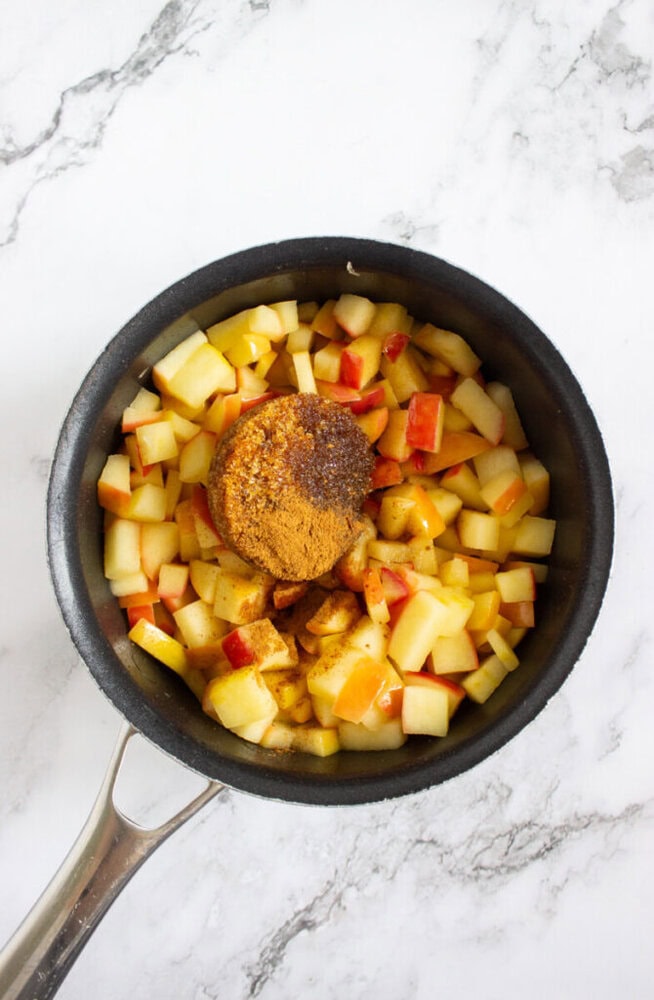 A saucepan filled with chopped apples, brown sugar, and ground cinnamon on a white marble surface.