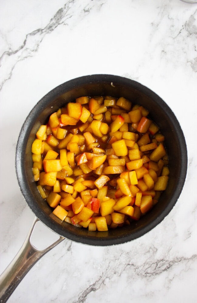 A saucepan filled with small diced apples cooked in a brown liquid sits on a white marble surface.