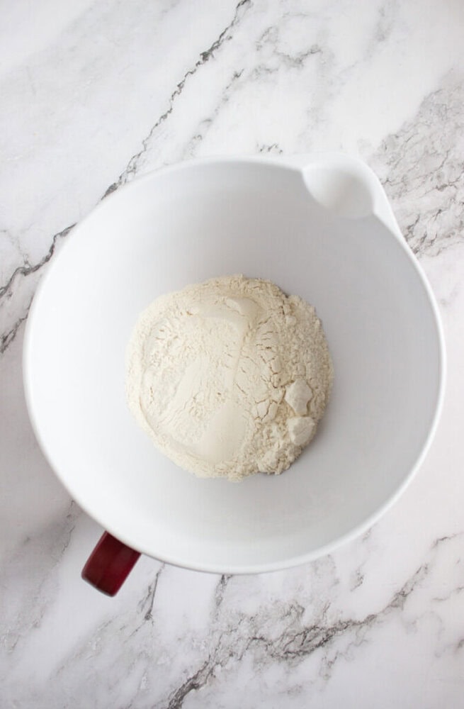 A white mixing bowl with a handle contains a mound of flour on a marble countertop.