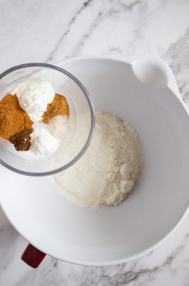 A white mixing bowl with flour inside sits on a marble surface. A measuring cup above the bowl contains spices and baking powder, about to be added to the flour.
