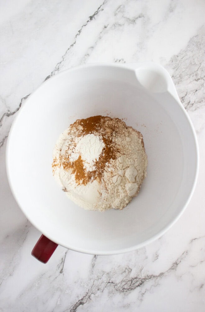 A white mixing bowl containing flour and cinnamon sits on a marble countertop.