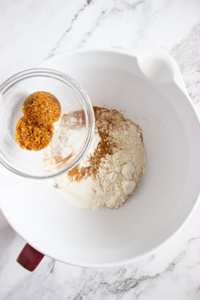 A glass bowl of brown sugar is being poured into a large white mixing bowl containing flour and spices on a marble surface.