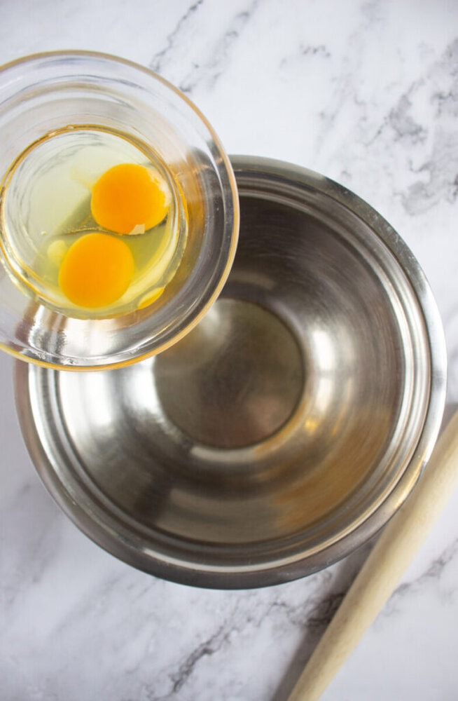 A glass bowl with two cracked eggs is held above an empty stainless steel mixing bowl on a marble countertop, alongside a wooden spoon.