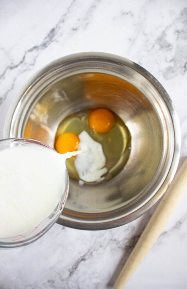 A metal bowl with two eggs and some milk being poured in, on a marble countertop with a wooden spoon beside it.