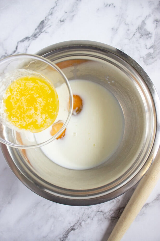 A glass bowl of melted butter is being poured into a metal mixing bowl containing milk and eggs on a marble countertop.