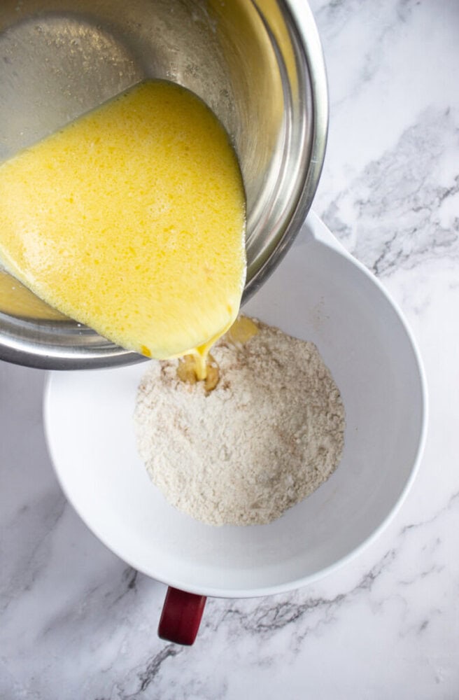 A metal bowl with beaten eggs is being poured into a white bowl containing flour on a marble countertop.