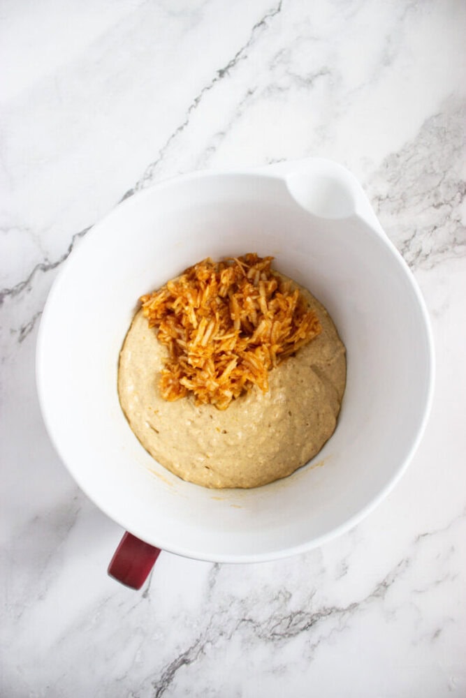 A white mixing bowl containing a thick beige batter topped with a mound of shredded, orange-colored ingredients on a marble surface.