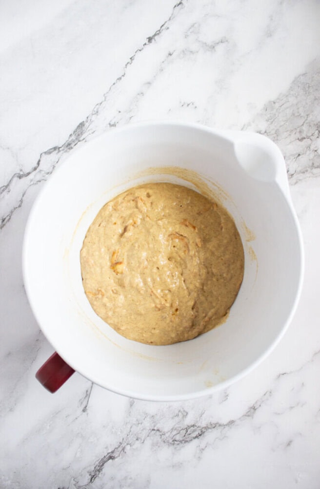 A white mixing bowl with light brown batter inside, placed on a white marble surface.