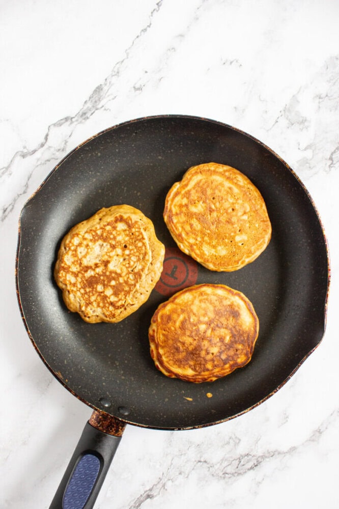 Three golden brown pancakes cooking in a black nonstick frying pan on a white marble surface.