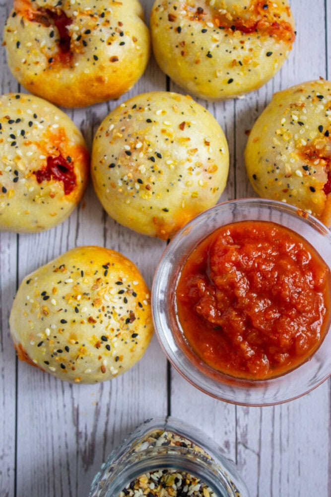 Six baked rolls topped with seasoning are arranged around a glass bowl of red marinara sauce on a wooden surface.