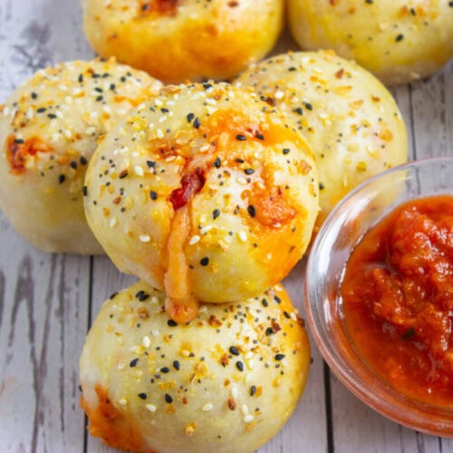 Baked dough balls topped with sesame and poppy seeds, some with visible melted cheese and tomato sauce, served with a bowl of red marinara sauce on a wooden surface.
