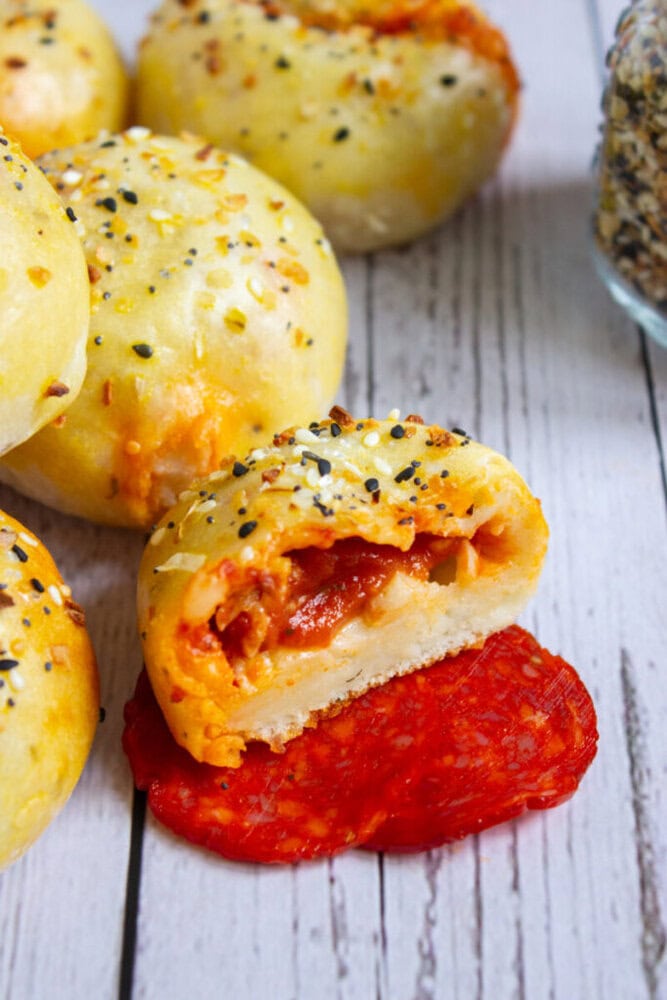 A batch of golden-brown, everything-seasoned pizza bites, one cut open to reveal melted cheese and pepperoni filling, on a white wooden surface.