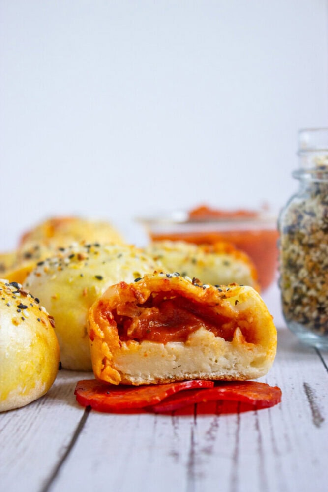 Close-up of a baked dough ball with a visible tomato and cheese filling, placed on sliced pepperoni, with more dough balls and jars of seasoning and sauce in the background.