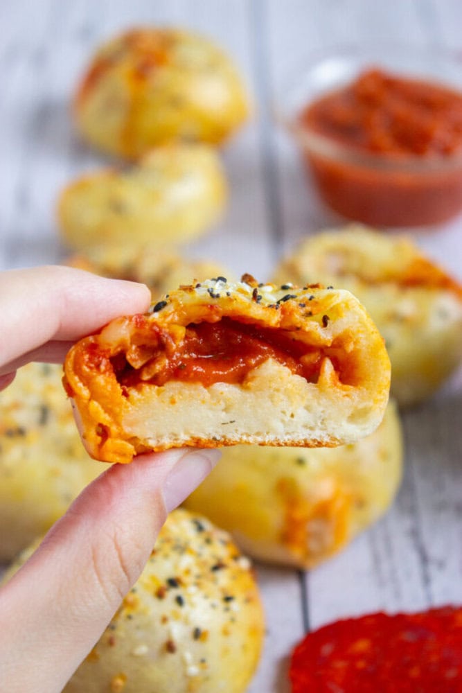 A hand holds a halved stuffed bread roll showing a tomato filling, with more rolls and a bowl of tomato sauce in the background.