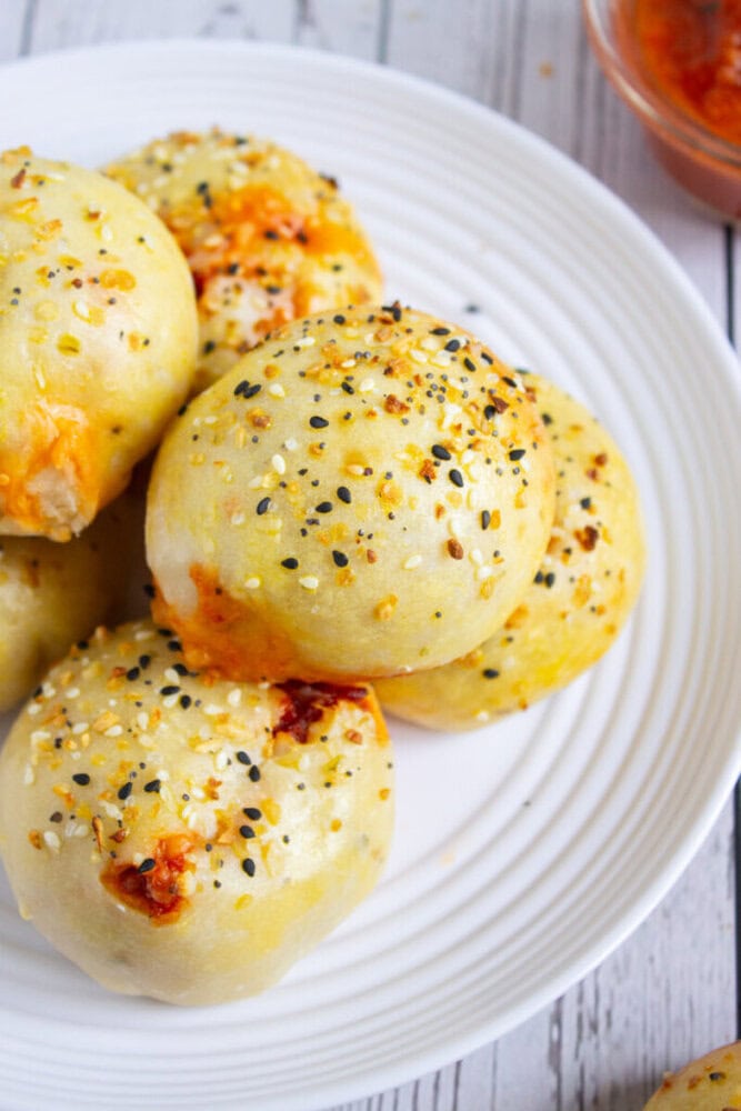 A white plate holds several round, golden-brown baked rolls topped with mixed seeds and bits of melted cheese, with a small bowl of sauce in the background.