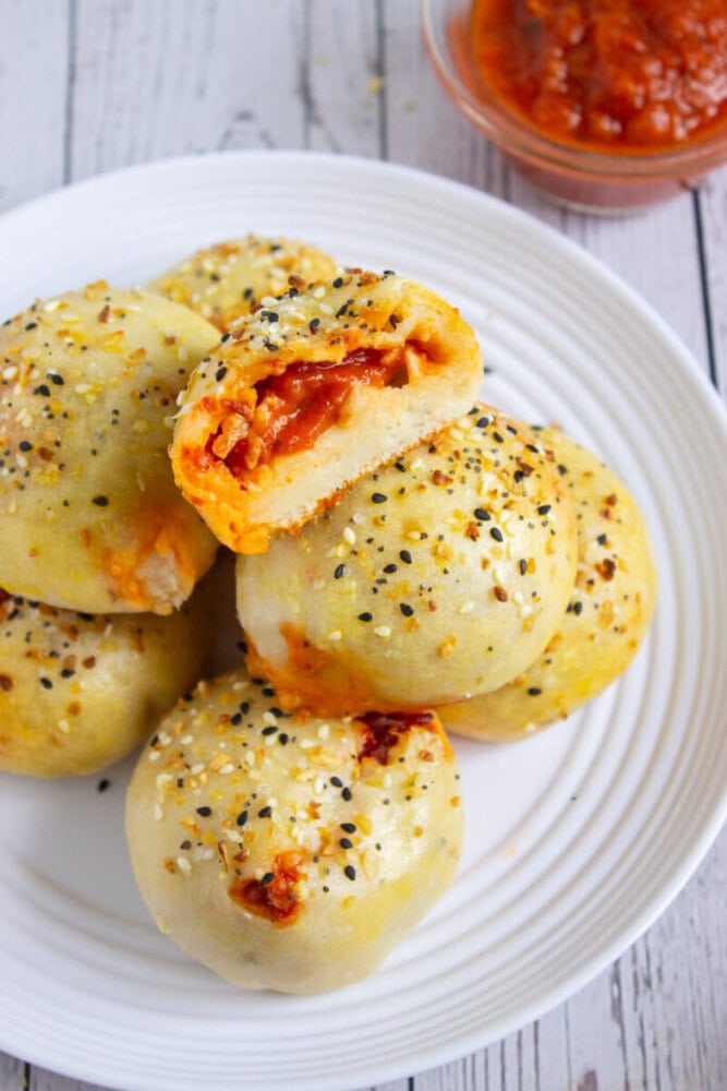 A plate of round, sesame-seed topped baked rolls filled with tomato sauce and cheese, with a bowl of red dipping sauce in the background.