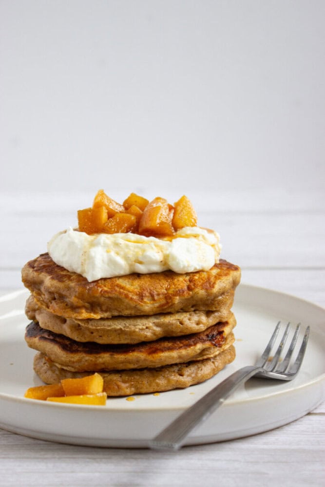 A stack of pancakes topped with whipped cream and diced peaches sits on a white plate with a fork beside it.