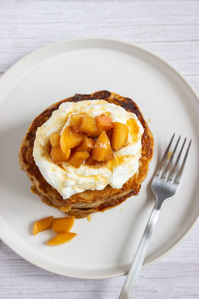 A stack of pancakes topped with whipped cream and diced caramelized apples on a white plate, with a fork beside the pancakes.