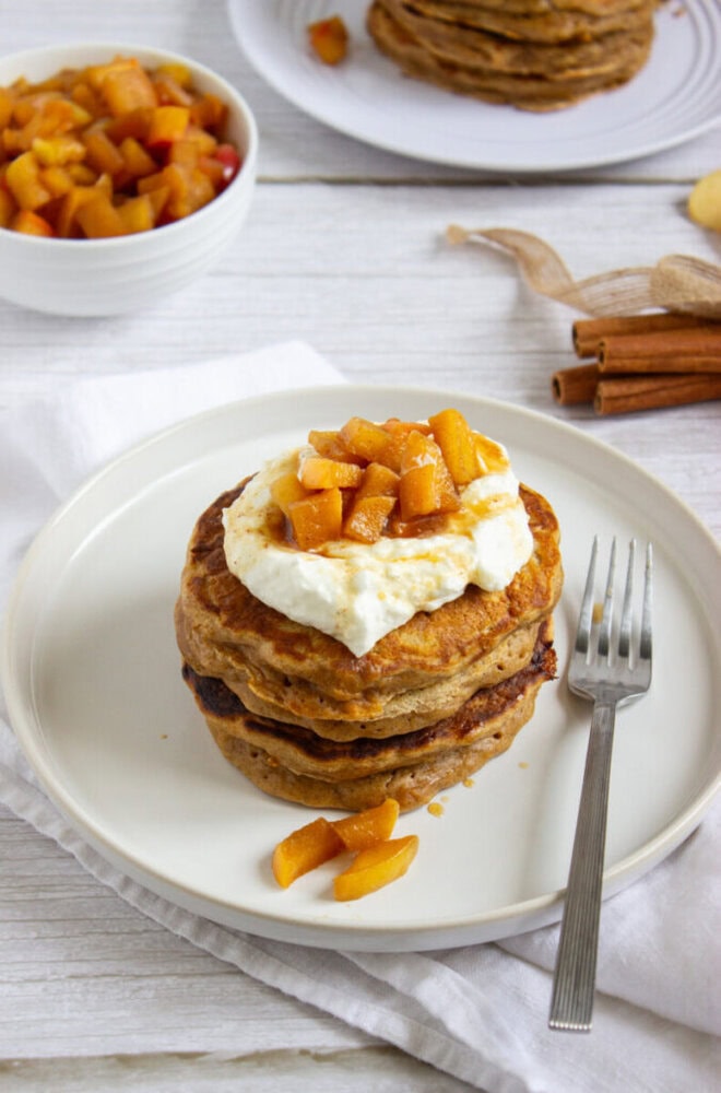 A stack of pancakes topped with whipped cream and diced cooked apples sits on a white plate with a fork, next to cinnamon sticks and a bowl of more apple topping.