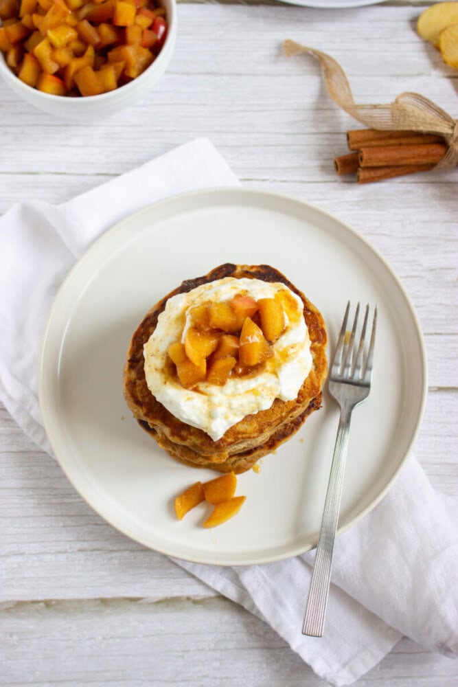 A stack of pancakes topped with whipped cream and diced peaches on a white plate with a fork, set on a white napkin.
