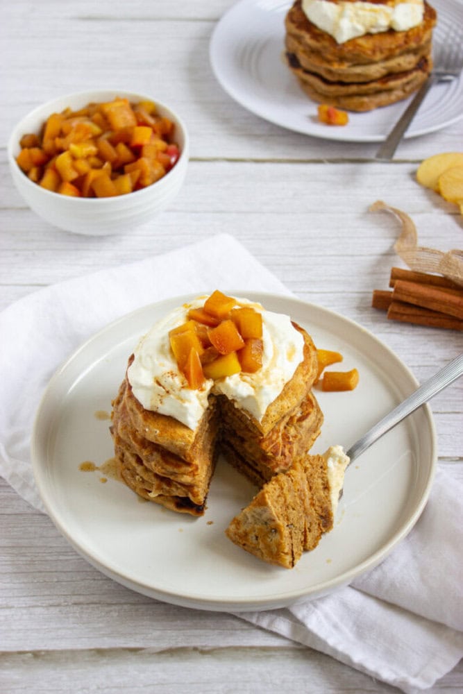 A stack of pancakes topped with cream and diced peaches on a white plate, with a slice cut out and a fork, and a bowl of diced peaches in the background.