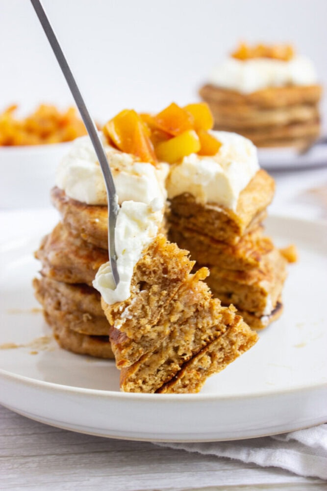A fork lifts a bite from a stack of pancakes topped with whipped cream and diced fruit on a white plate.