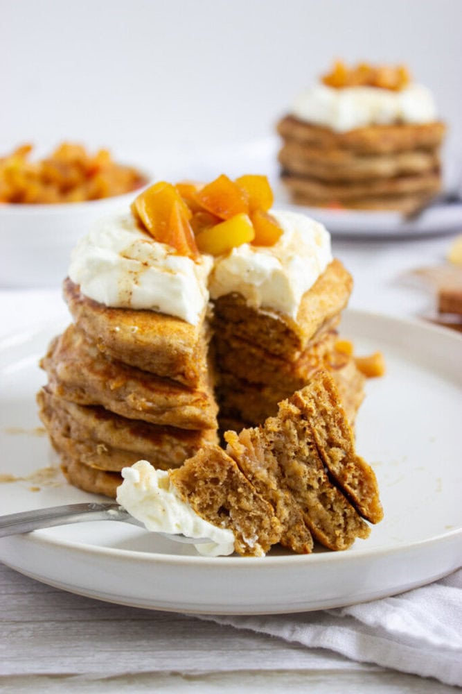 A stack of pancakes topped with whipped cream and diced peaches on a white plate, with a fork holding a cut piece in the foreground.