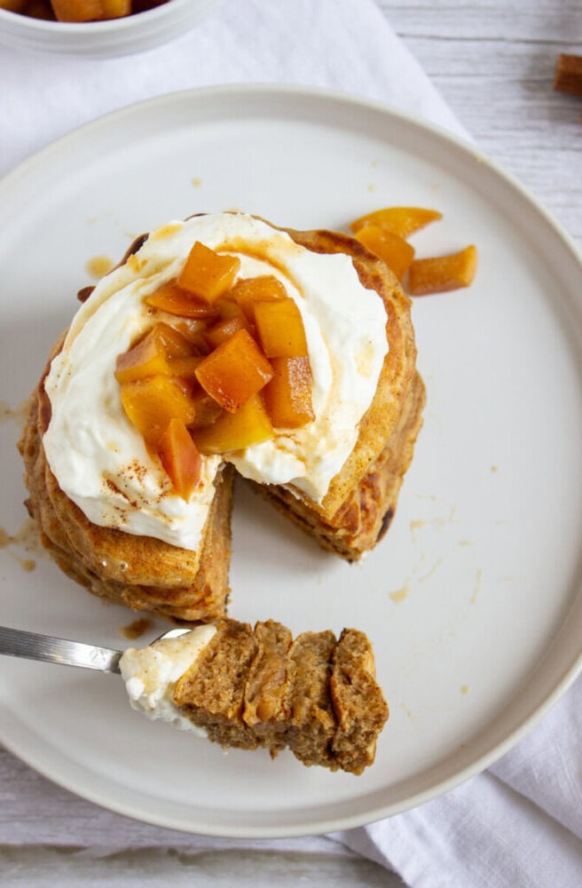 A plate with a stack of pancakes topped with whipped cream and diced fruit, with a fork holding a slice of the pancakes.